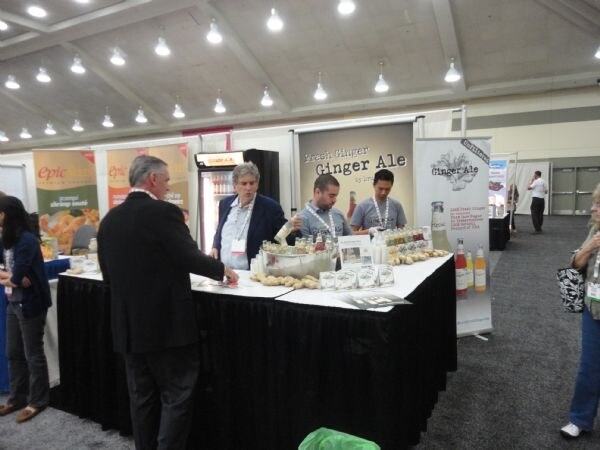 Several people stand at a black-skirted counter under a Fresh Ginger Ale banner, with bottles on display. An Epic booth is visible to the left.