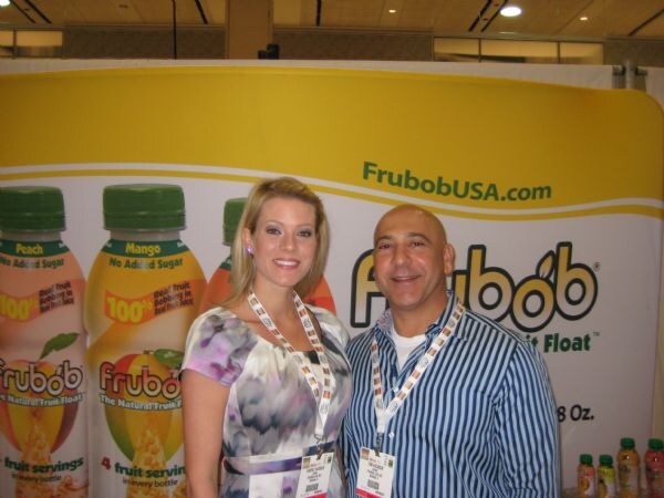 A smiling woman in a patterned dress and a man in a blue striped shirt, both wearing lanyards, stand in front of a Frubob banner showing peach and mango fruit drink bottles.