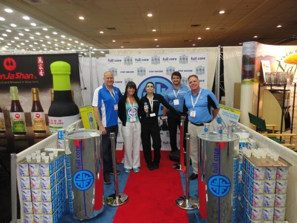 Five people stand on a red carpet in a Full Core booth with blue Full Core banners, silver display tubs and stacked cartons of water. A Jin Ja Shan display is at left.