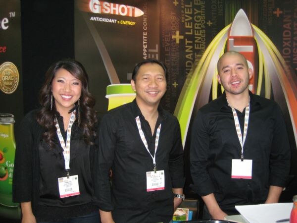 Two men and a woman in black clothing and lanyards stand smiling in front of a dark G Shot antioxidant and energy banner with a rocket graphic.