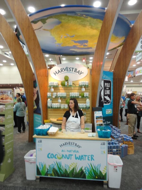 A woman in a white apron stands at a Harvest Bay all-natural coconut water booth with a globe overhead. A Pole and Line banner stands to the right.