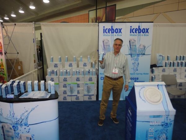A man in a striped shirt and khaki pants holds up an icebox carton in a booth with icebox Water in a Box banners and stacked cartons of boxed water.