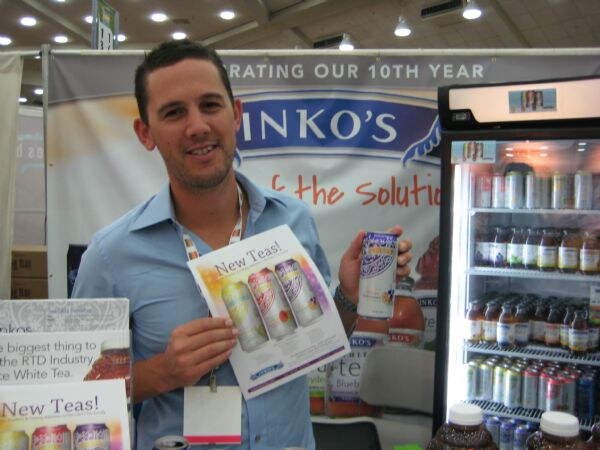 A smiling man in a light blue shirt holds up a can and a flyer reading New Teas! in front of an Inko's banner and a refrigerator stocked with bottled and canned iced tea.