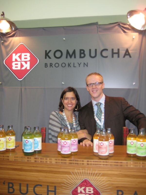 A smiling woman and a man with glasses, a striped jacket and green tie stand behind a wooden counter lined with bottles, in front of a gray Kombucha Brooklyn banner with a red KB logo.