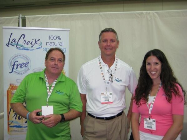 Three smiling people wearing show badges stand together in front of a LaCroix banner reading 100% natural sparkling water. Two wear LaCroix polo shirts, one green and one white, and one wears pink.