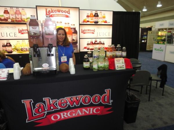 A woman in a blue top stands behind a black-draped Lakewood Organic counter with a juice dispenser, bottles of juice and coconuts, in front of backlit juice displays. A Native sign is at right.