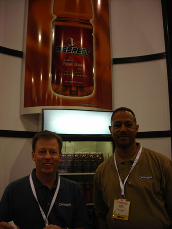 Two men in polo shirts with lanyards stand below a large backlit Respect bottle display above a lit refrigerated cooler holding bottles.