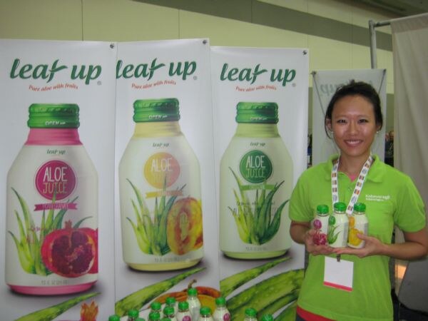 A smiling woman in a green polo holds three small bottles of aloe juice beside tall Leaf Up banners showing pomegranate, citrus and plain aloe juice flavors.