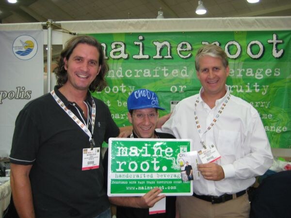 Three men pose in front of a green Maine Root banner. The man in the middle wears a blue cap and holds a green Maine Root sign. The other two wear show badges.