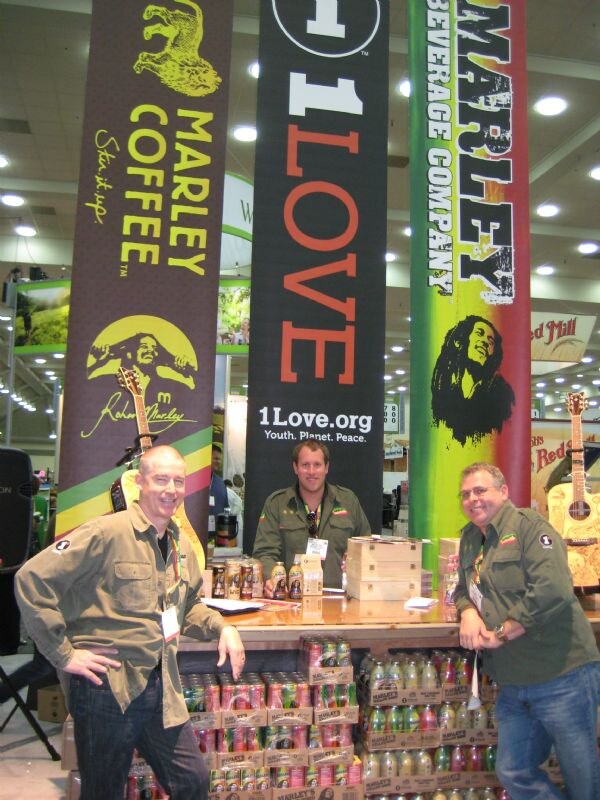 Three men in olive green shirts stand around a wooden counter stacked with canned and bottled drinks, beneath tall banners for Marley Coffee, 1Love and Marley Beverage Company.