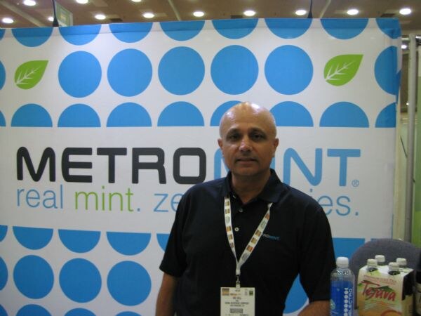 A bald man in a dark polo with a show badge stands in front of a blue and white Metromint banner. A water bottle and a Tejava carton sit at the lower right.