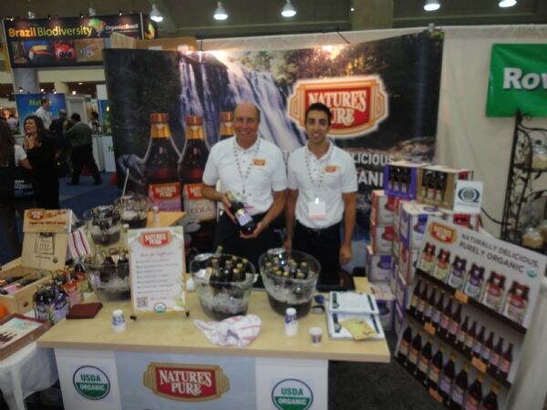 Two men in white polo shirts stand behind a table with bowls of iced bottles at the Nature's Pure booth. Cases and shelves of bottles and USDA Organic signs surround them.