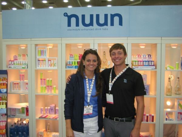 A woman in a navy jacket and a man in a black polo stand in front of lit shelves of nuun electrolyte tab tubes beneath a blue nuun sign reading electrolyte enhanced drink tabs.