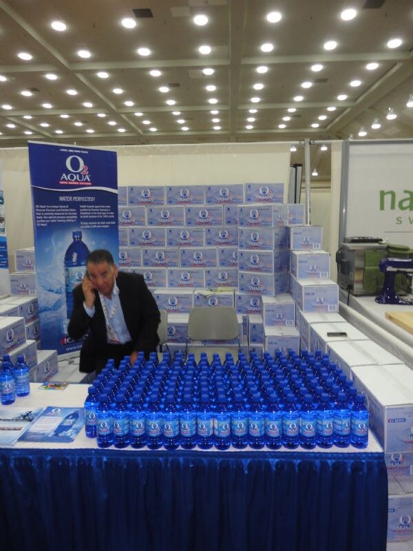 A man in a dark jacket talks on a phone behind a blue-draped table covered with rows of O2 Aqua water bottles. Stacked O2 Aqua cases and a banner stand behind him.