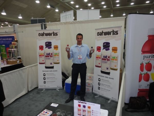 A man in a light blue shirt holds up two small bottles between two tall Oatworks banners showing bottles and the slogan because oats work. A Purity Organic banner is at right.