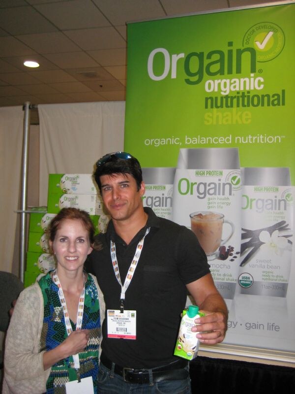 A woman in a green patterned top and a man in a black shirt with sunglasses on his head pose in front of a green Orgain organic nutritional shake banner. The man holds a small Orgain carton.