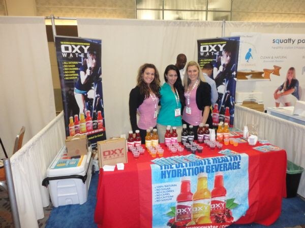 Three women stand behind a red-covered table of OXY Water bottles and sample cups, with OXY Water banners behind them. Two men stand in the background.