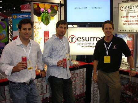 Three men with badges stand in front of a Resurrect Daily Detox and Anti-Hangover display; two hold red cans, and cases of product are stacked behind them.