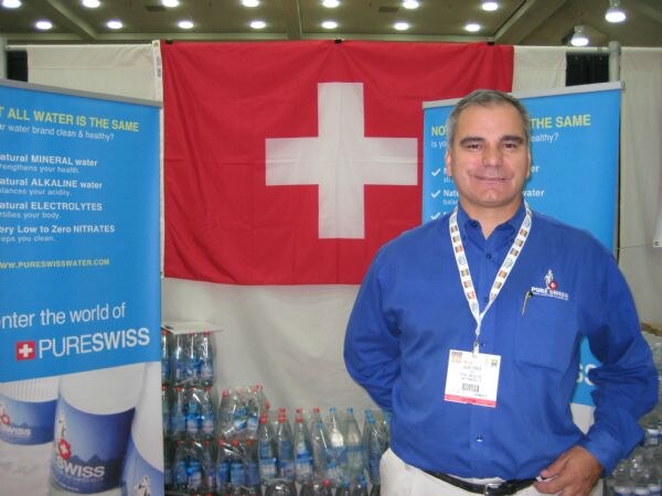 A man in a blue PureSwiss shirt and lanyard stands in front of a Swiss flag, with blue PureSwiss banners and stacked bottled water beside him.