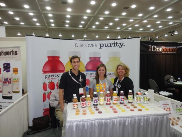 A man and two women stand behind a white-draped table of Purity Organic juice bottles and sample cups, in front of a purity organic banner. An OneSource sign is at right.