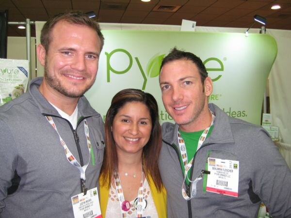 Close-up of two men in gray jackets and a woman in a yellow cardigan, all wearing badges, smiling in front of a green Pyure sign. Benjamin Fleischer of Pyure Brands LLC is on the right.