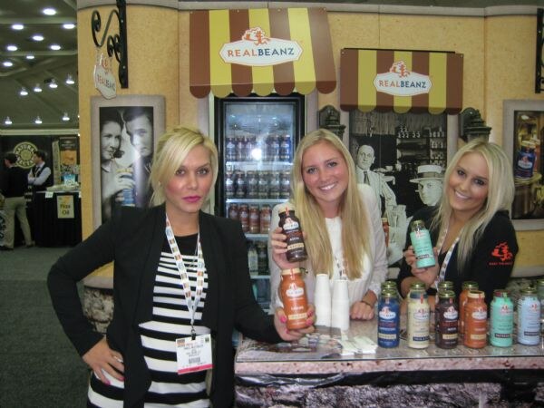 Three blonde women pose at a Real Beanz booth with striped awnings, holding bottled coffee drinks. More bottles sit on the counter and in a refrigerator behind them.