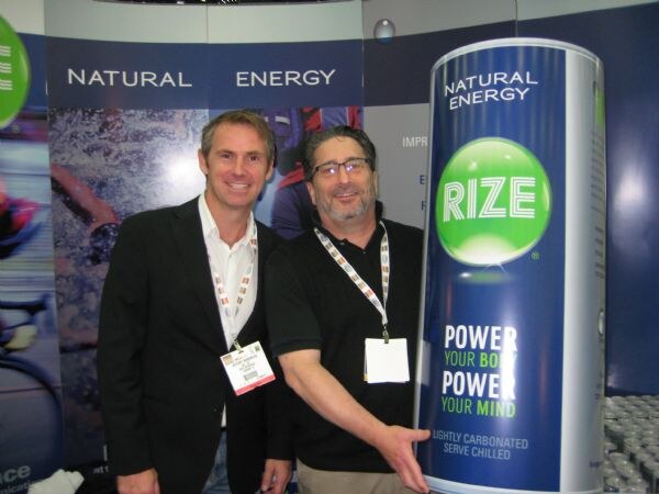 Two men in dark clothing and lanyards stand beside a large Rize natural energy can display. One man wears glasses and gestures toward the can.