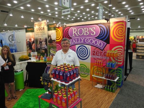 A smiling man in a white polo stands behind a display of purple and orange bottles in a colorful Rob's Really Good booth with Zero signage and shelves of drinks.
