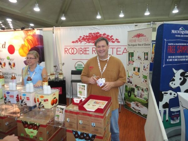 A smiling man in a brown shirt holds a bottle at the Roibee Red Tea booth, while a woman stands at left behind sample dispensers. A Mountain High banner is at right.