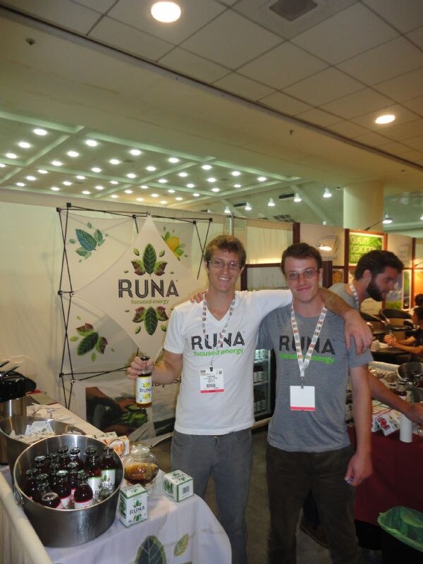Two men in Runa t-shirts and badge lanyards stand with arms around each other at the Runa booth; one holds a bottle, and a tub of bottled drinks sits on the table in front.