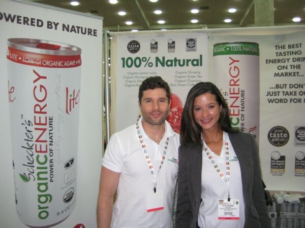A man and a woman in white shirts and badge lanyards stand in front of large Schecter's Organic Energy banners showing a can and the text 100% Natural.