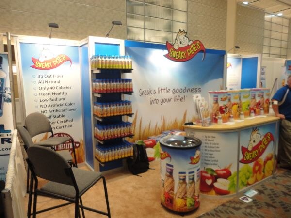 Sneaky Pete's booth with blue graphic walls, a shelf of bottled drinks, a counter with product displays, a branded round bin and gray chairs. The slogan reads Sneak a little goodness into your life.