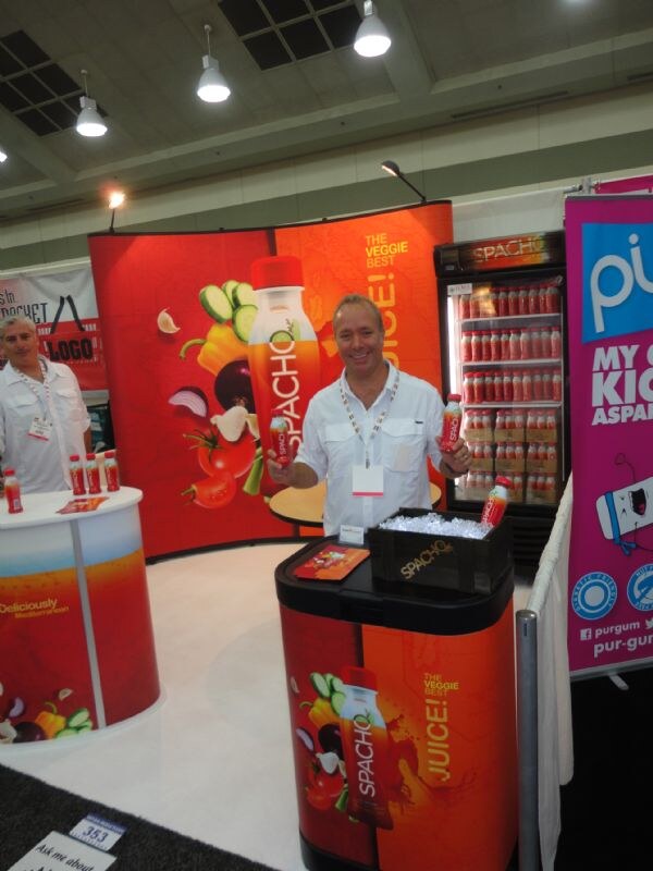A smiling man in a white shirt holds a Spacho bottle behind a counter at a red Spacho booth. A refrigerator of bottles is behind him, and a Pur banner is at right.