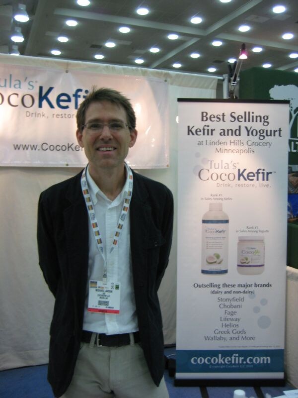 A smiling man with glasses, a dark jacket and a lanyard stands in front of a Tula's CocoKefir banner and a standing sign reading Best Selling Kefir and Yogurt.