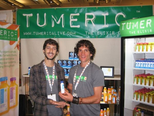 Two smiling men in gray shirts and lanyards stand in front of a green Tumeric banner. One holds a small white bottle, and refrigerated shelves of bottled drinks are at right.
