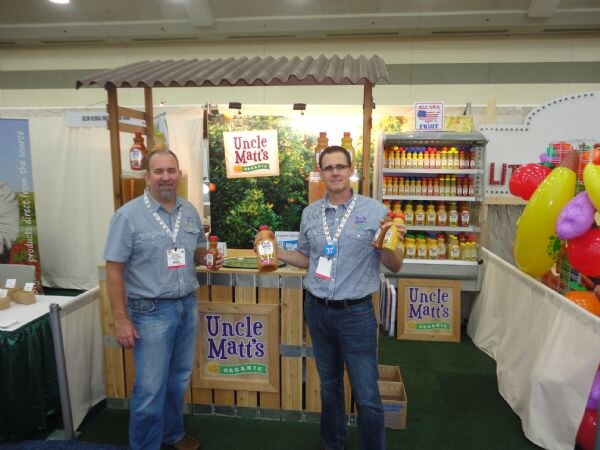 Two men in blue work shirts hold bottles of orange juice in front of the Uncle Matt's Organic booth, which has a wooden counter, signs and a shelf of bottles.