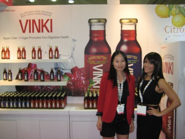 Two women stand in front of Vinki displays showing bottles of apple cider vinegar juice beverage on shelves and large bottle graphics. One wears a red jacket and the other a black dress.