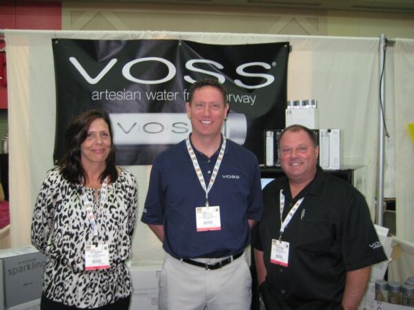 A woman and two men wearing badges stand together in front of a black Voss banner reading artesian water from Norway. Cases of sparkling water are at lower left.
