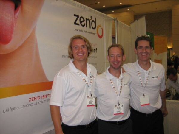 Three smiling men in white polo shirts and badge lanyards stand together in front of a Zen'd banner.