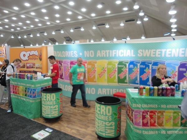Zevia booth with a banner of colorful soda cans and the text zero calories, green barrels and a table of cans. A Qrunch Burgers sign is at left; several attendees and staff are present.