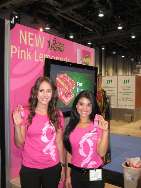 Two women in pink shirts hold small bottles in front of a pink 5-hour ENERGY Pink Lemonade sign. A JTI sign is visible in the background.