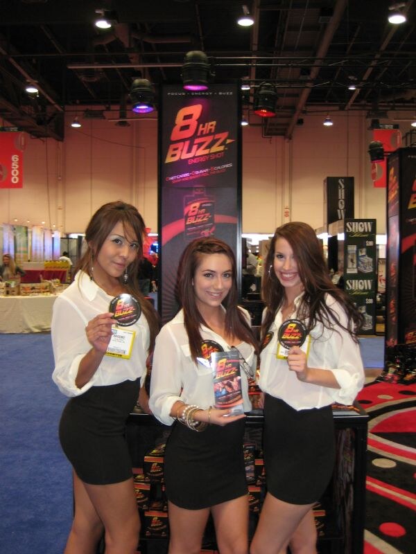 Three smiling women in white blouses and black skirts hold 8 Hour Buzz round badges and a product package in front of an 8HR Buzz Energy Shot banner.