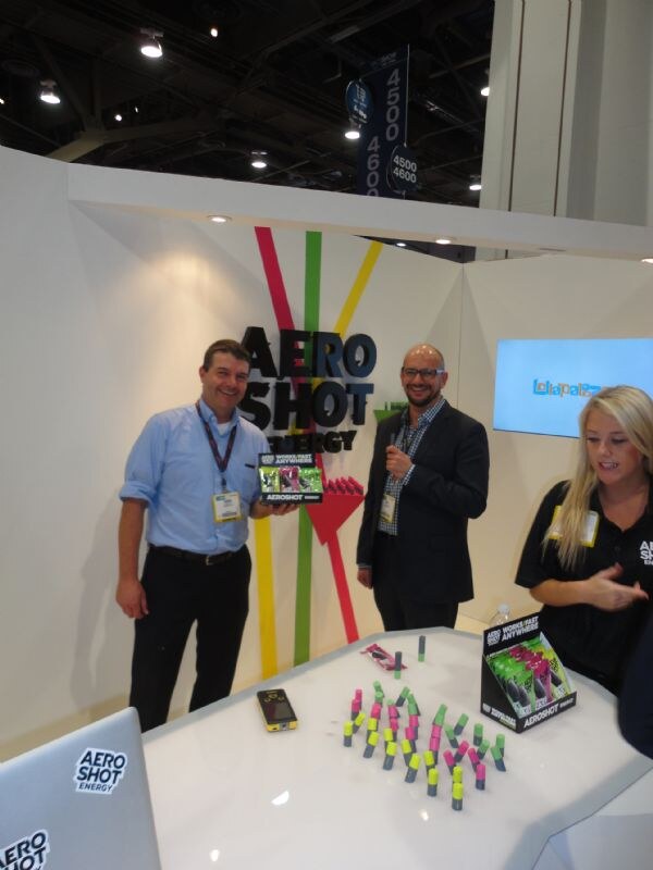 Two men and a blonde woman at an AeroShot Energy booth; one man holds an AeroShot box, and a table in front is covered with pink and green AeroShot canisters.
