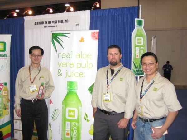 Three men in tan shirts and lanyards stand at the ALO Drink by SPI West Port booth beside a banner reading real aloe vera pulp & juice; one holds an Alo bottle.