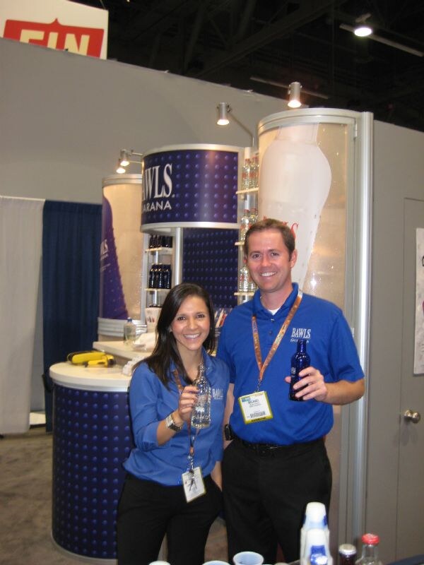 A smiling woman and man in blue Bawls shirts hold glass Bawls bottles in front of a booth with blue dotted display towers bearing the Bawls Guarana name.