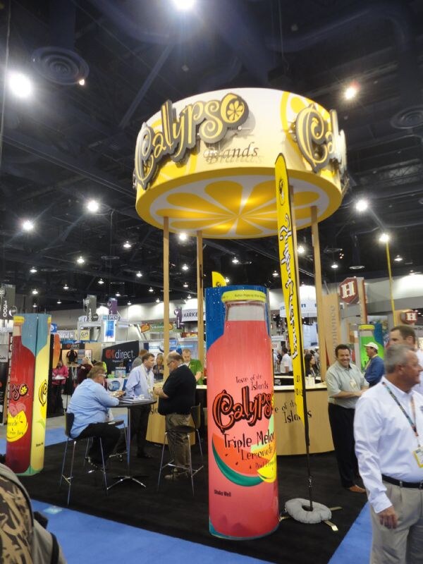 The Calypso Brands booth has a large lemon-slice canopy and tall Calypso lemonade bottle displays, with attendees talking at tables and standing nearby.