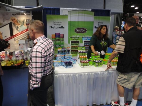 A woman in a green shirt stands behind a table of Celestial Seasonings natural shots while two men look at the display in front of green banners.