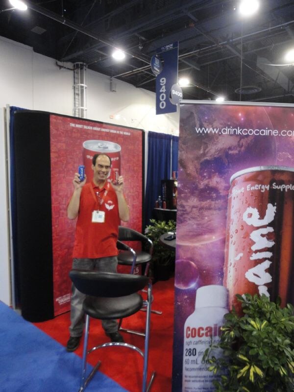 A man in a red shirt holds up two small cans in front of a red Cocaine backdrop; a banner showing a can and the drinkcocaine.com address is at right.