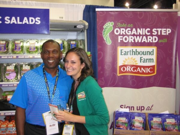 A smiling man in a blue shirt and a woman in a green Earthbound Farm jacket stand in front of a purple Earthbound Farm Organic banner and a refrigerated salad case.