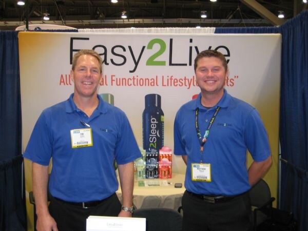 Two smiling men in blue polo shirts and exhibitor badges stand in front of an Easy2Live booth backdrop showing a tall 2Sleep bottle and other products on a table.